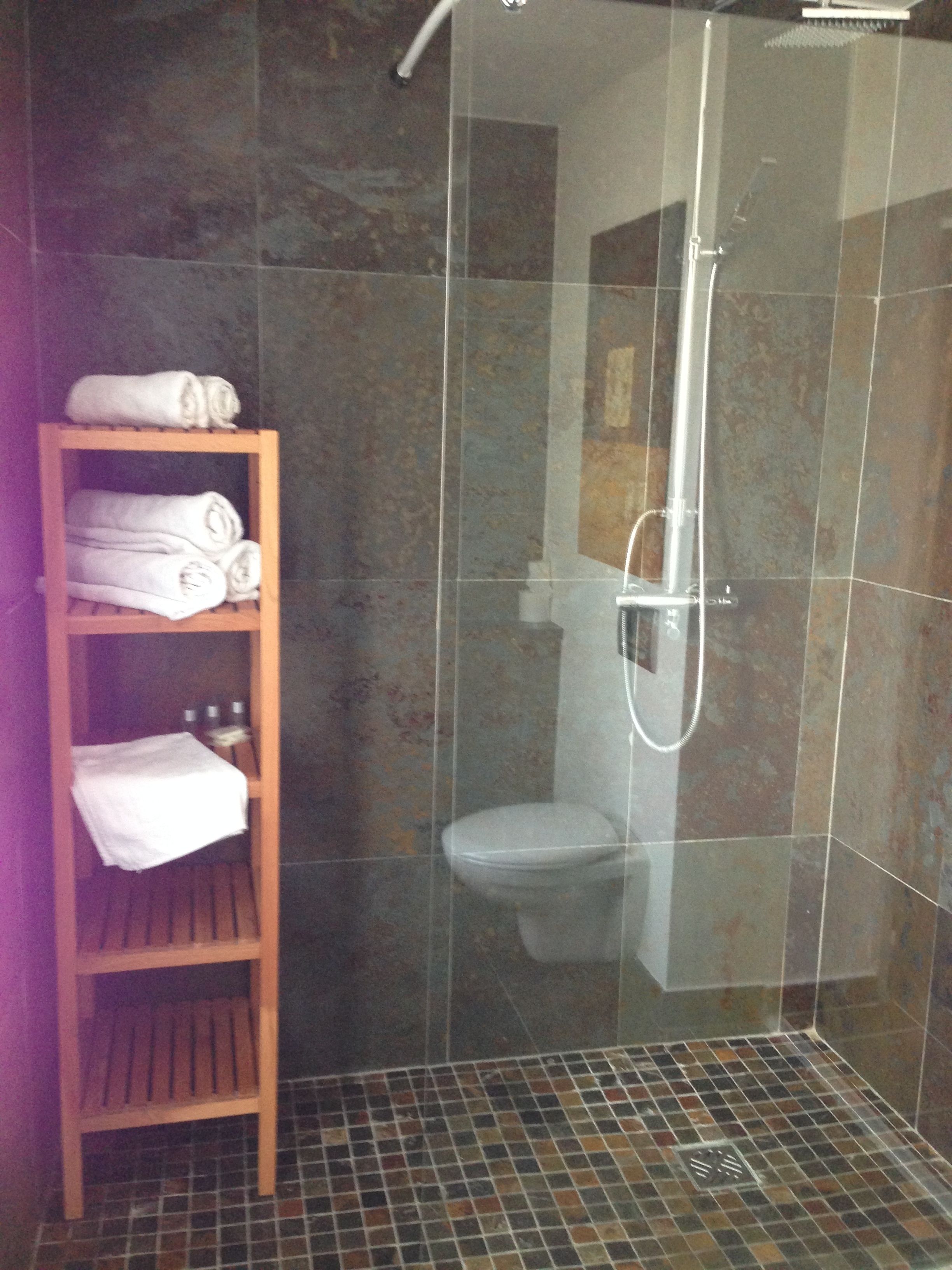 Cabina de ducha con paredes de vidrio, un cabezal de ducha de mano, un inodoro y un estante de madera con toallas.