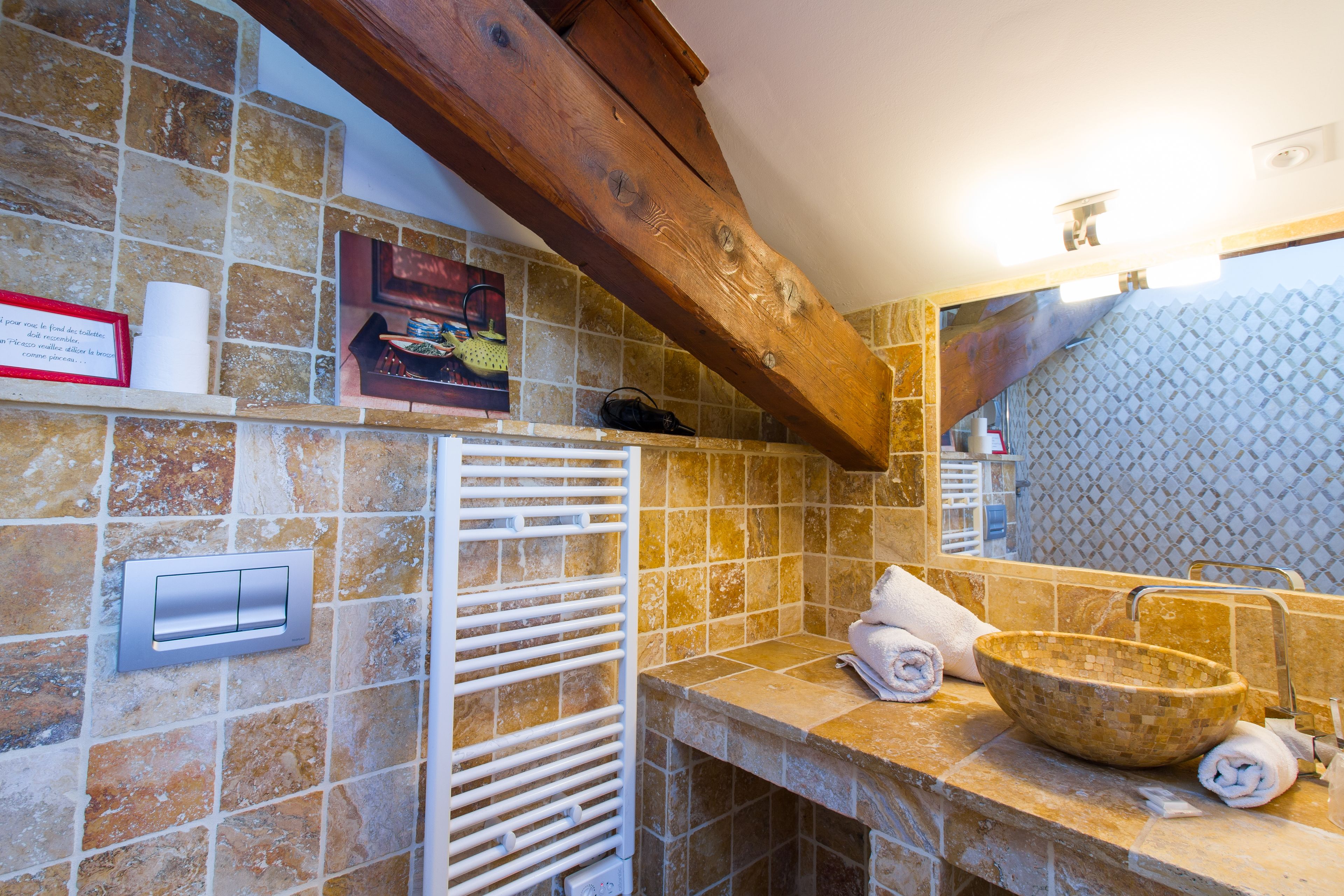 Une salle de bain avec des carreaux de pierre, une poutre en bois, un porte-serviettes et un lavabo surmonté d’une vasque en mosaïque.