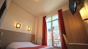 A hotel room with a bed, a window with red curtains, a wall-mounted TV, and a radiator. - Hôtel de la Cathédrale (Reims)