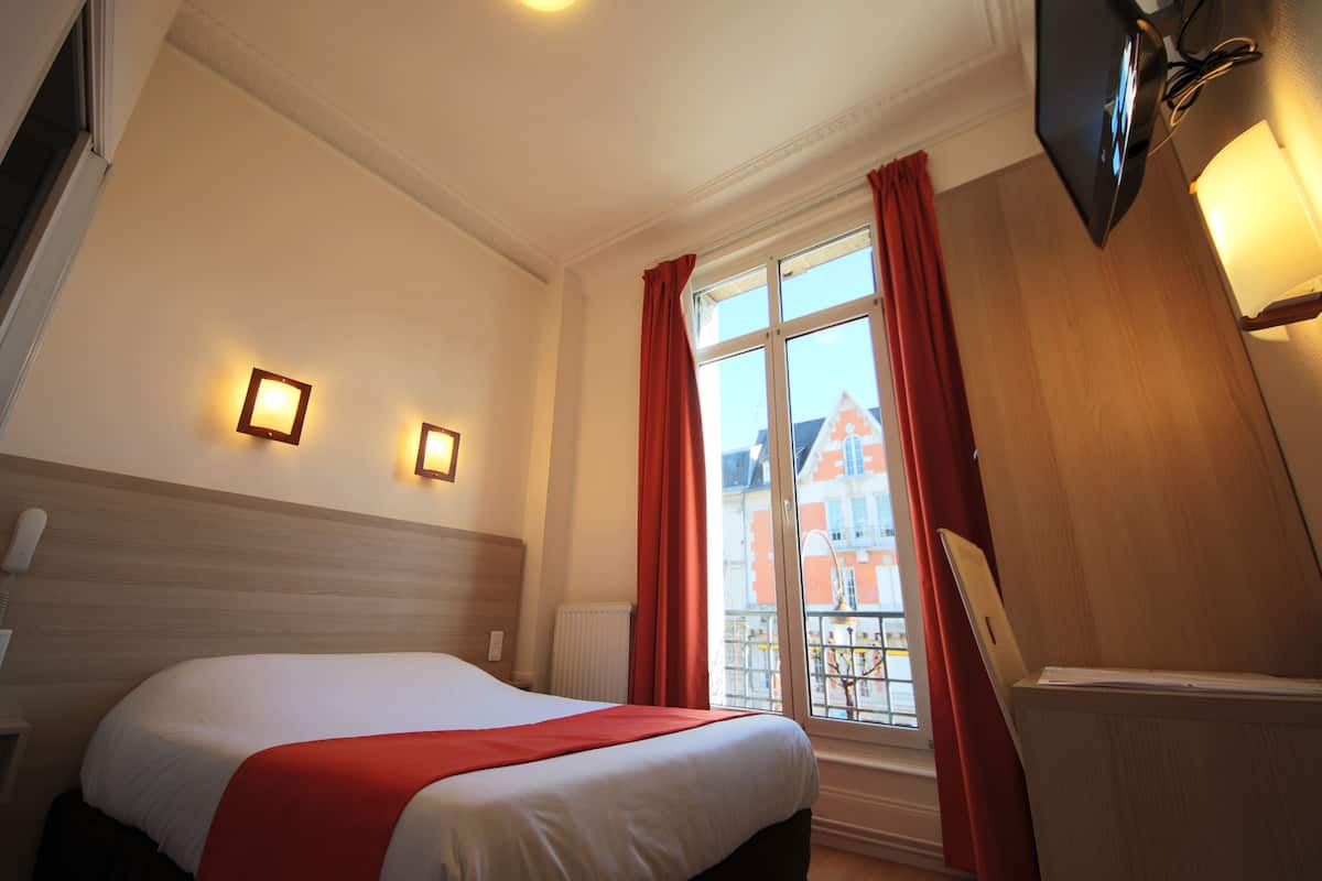 A hotel room with a bed, a window with red curtains, a wall-mounted TV, and a radiator.