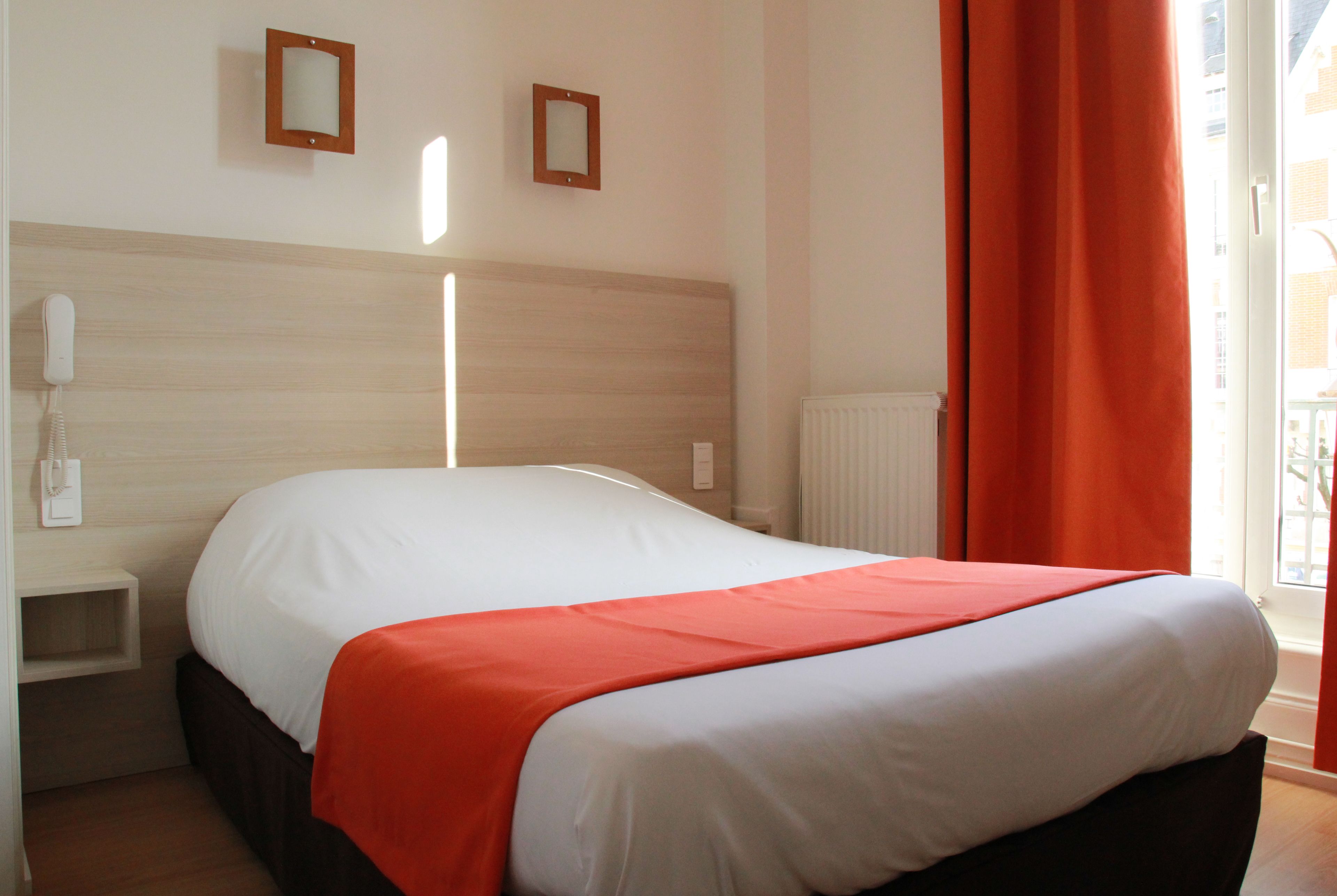 Un lit bien fait, avec une couvre-lit blanche et orange, une tête de lit en bois et un téléphone sur la table de chevet.