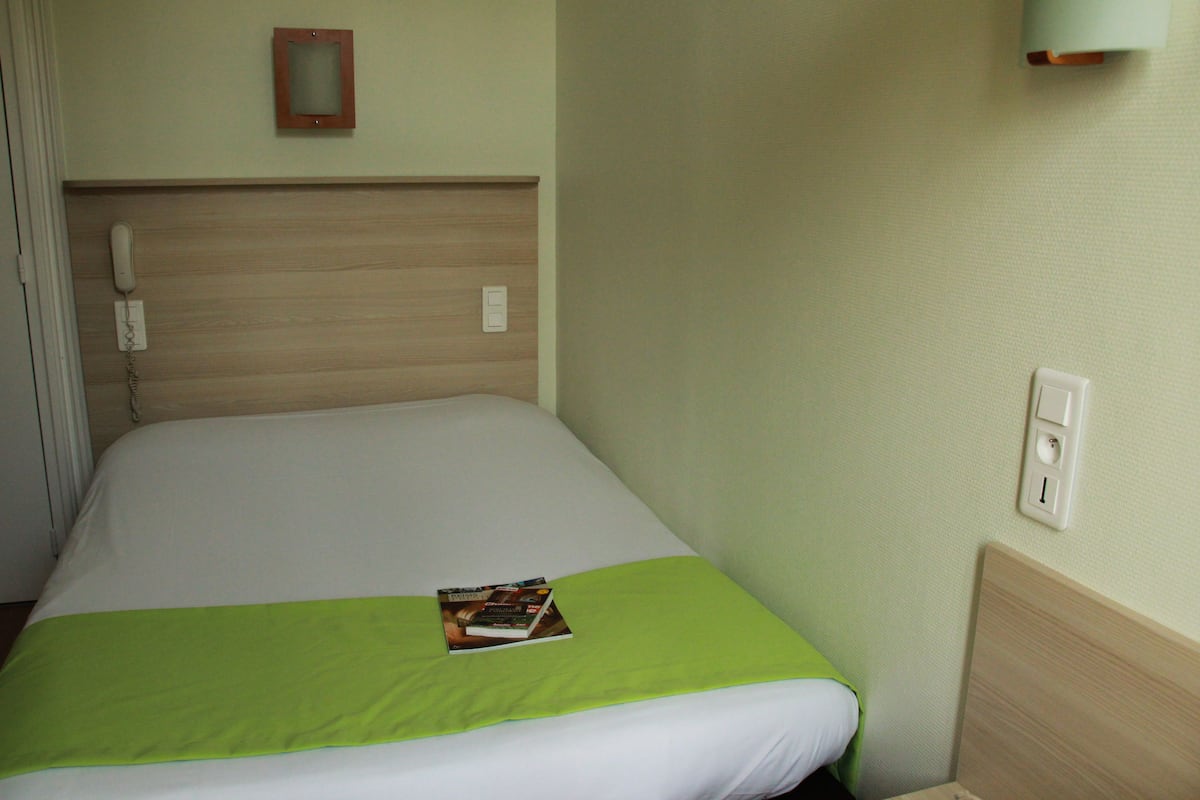 A hotel room with a bed, a bedside telephone, a light switch, and a framed picture on the wall.