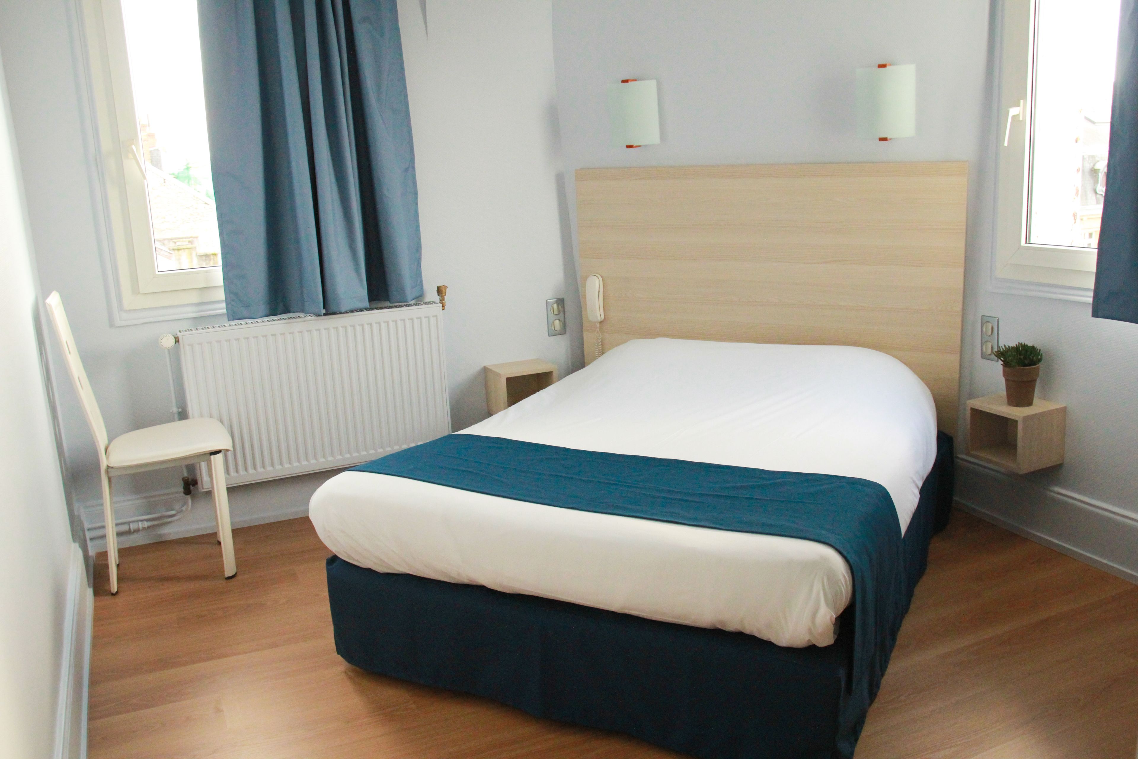 A bedroom with a wooden headboard, a white bed with a teal bedspread, a white chair, a small bedside table with a plant, and a window with blue curtains.