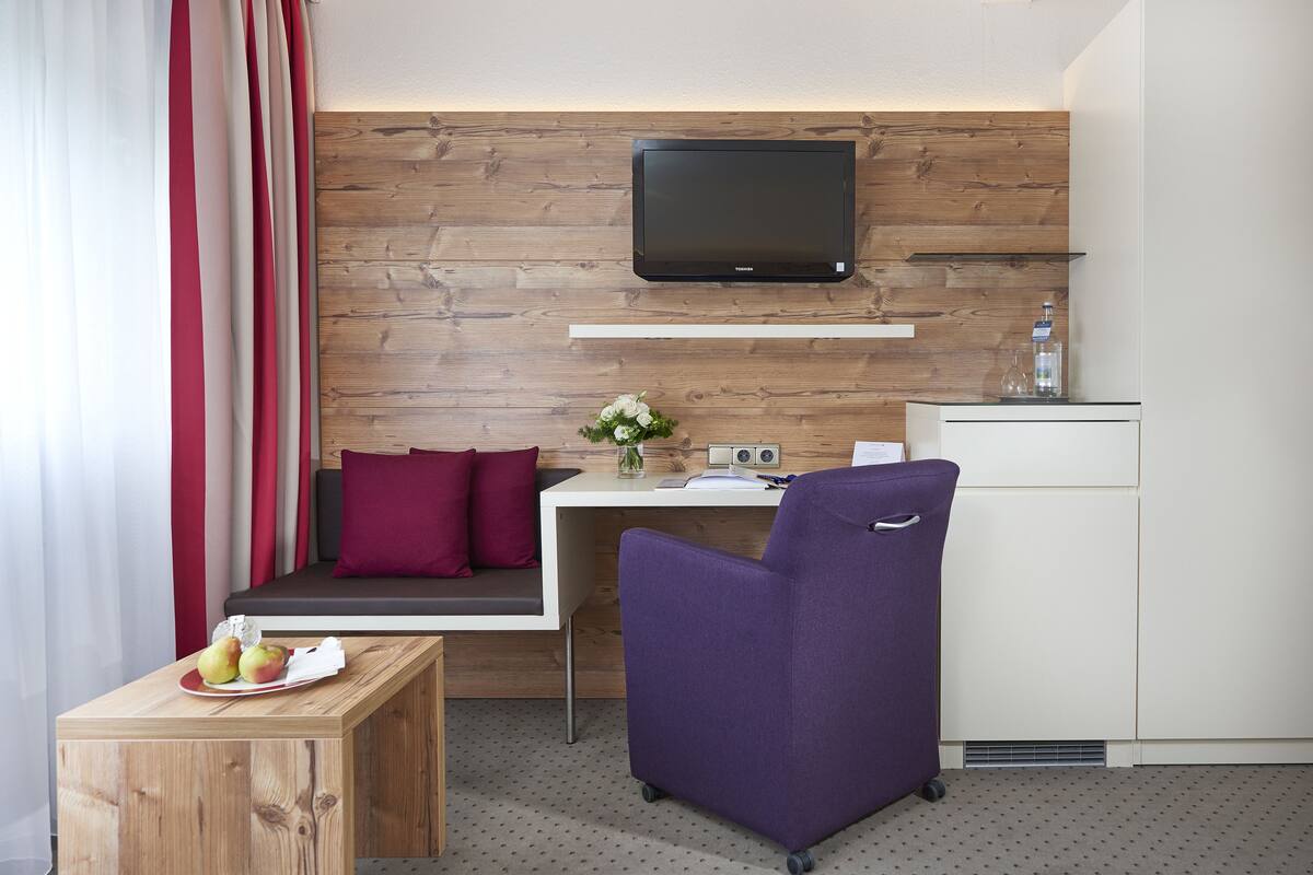 Ein modernes Hotelzimmer mit einer Holzwand, einem flachbildfernseher über einem Schreibtisch, einem violetten Sessel, einem kleinen Tisch mit Äpfeln und einer Couch mit roten Kissen.