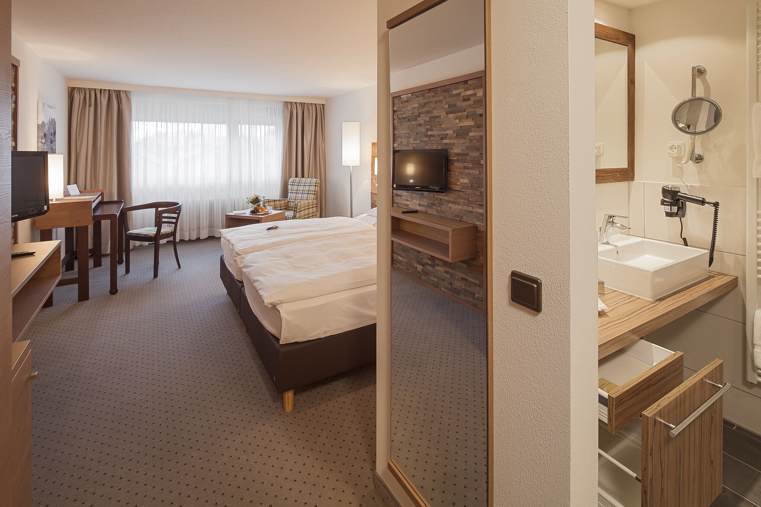 Ein Hotelzimmer mit Bett, Schreibtisch mit Stuhl, Fernseher, Bad mit Waschbecken und Spiegel sowie einer Dusche.