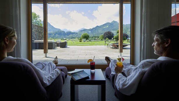 A room with a large window offering a view of a mountainous landscape. There is a table with a drink and a book. - Schüle's Gesundheitsresort & Spa - Erwachsenenhotel (Oberstdorf)