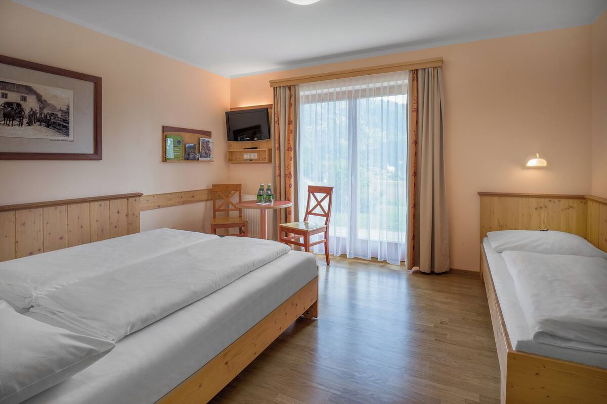 A hotel room with two single beds, a wooden headboard, a television, a small table with chairs, and a window with curtains.