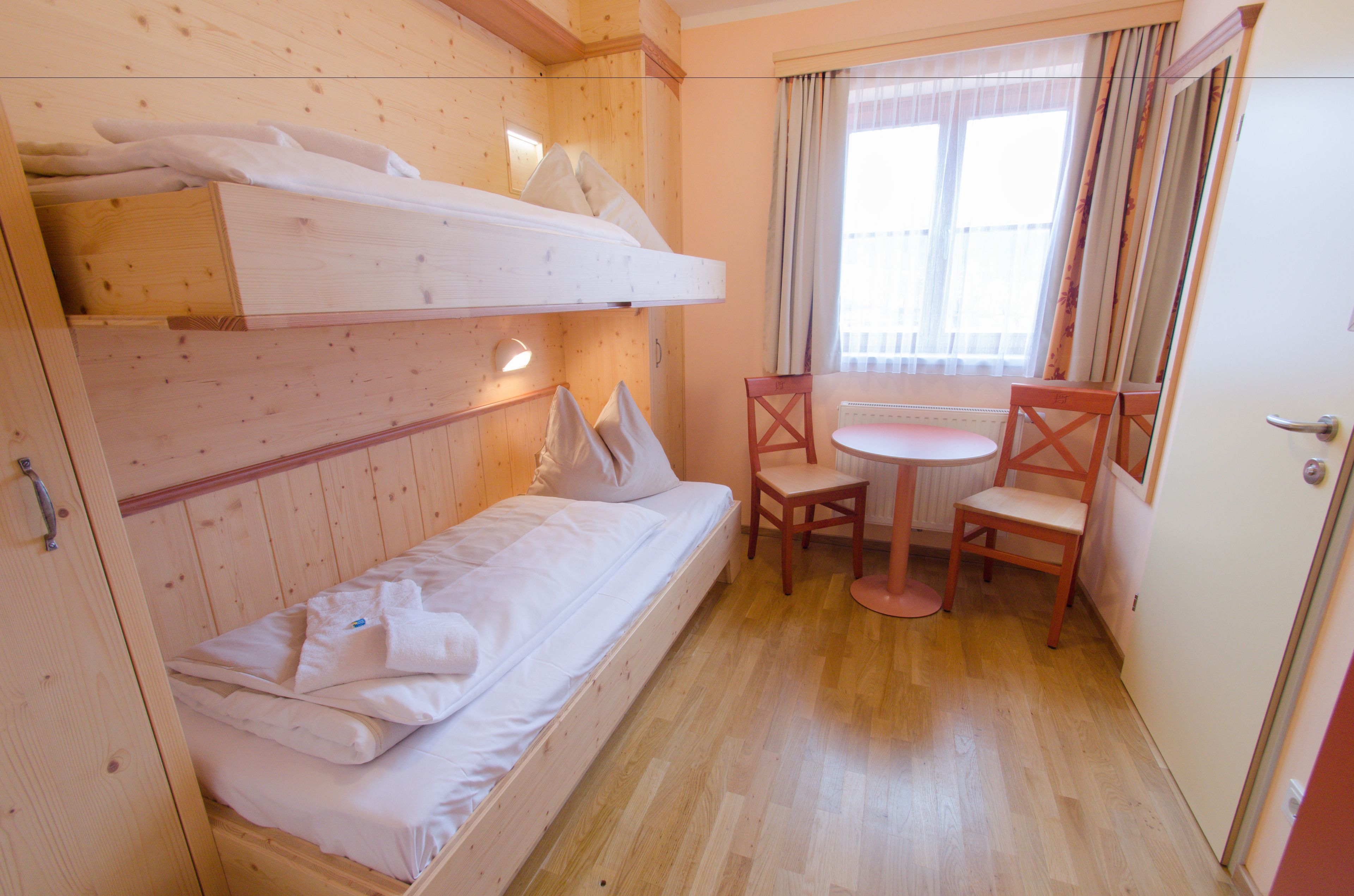 Une petite pièce agréable, dotée d’un lit superposé, d’une chaise, d’une petite table et d’une fenêtre avec des rideaux.