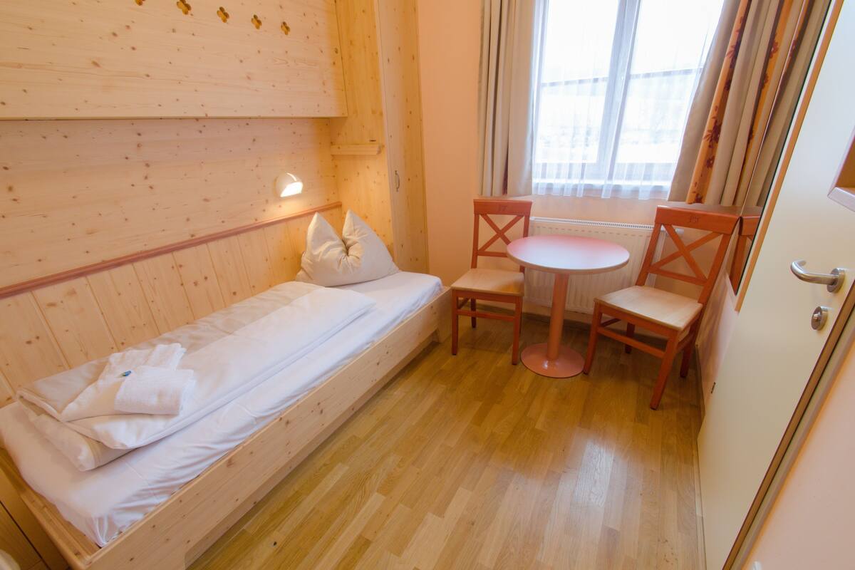 A small room with a single bed, a wooden chair, a small table, and a window with curtains.