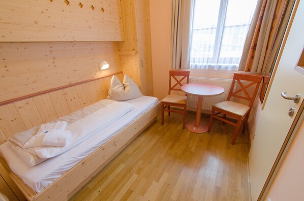 Una habitación pequeña con una cama individual, una silla de madera, una mesita y una ventana con cortinas.
