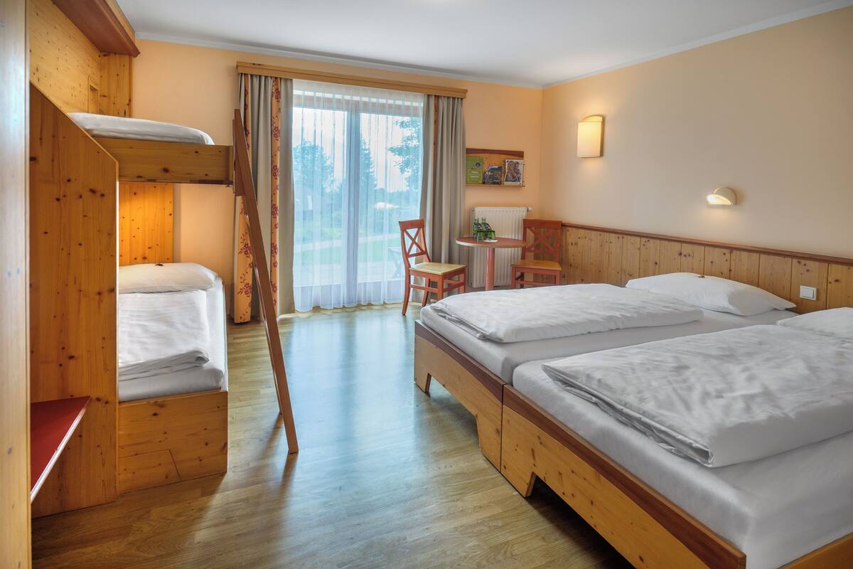 A hotel room with a bunk bed, a desk with a chair, a small table, and a view of the outdoors through the window.