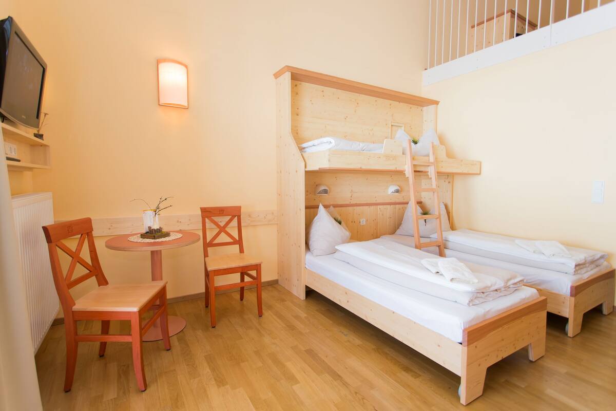 A room with two wooden bunk beds, a small table with a chair, a TV, and a wall-mounted light.
