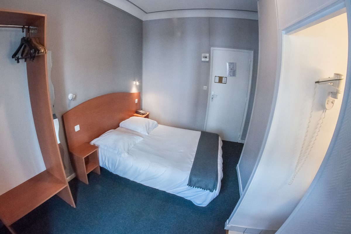 A hotel room with a bed, a wooden wardrobe, a bedside table, and a door.