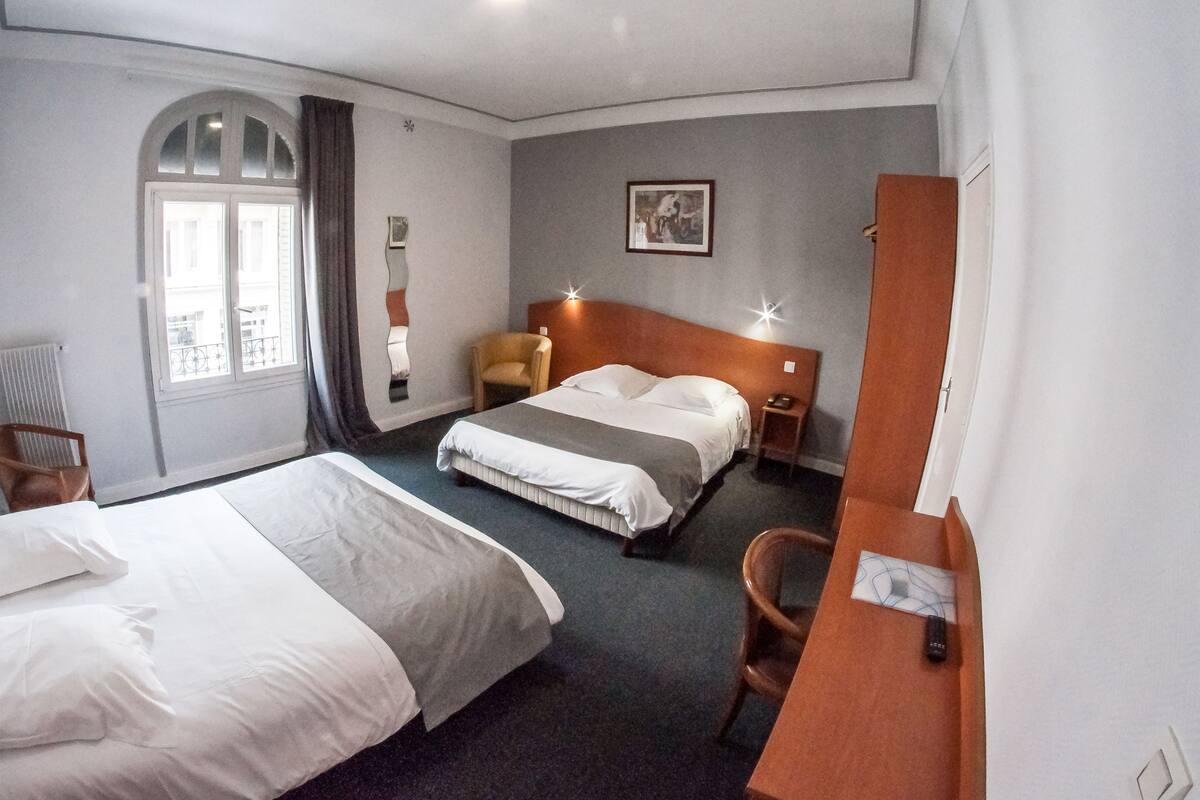 A hotel room with a large bed, a desk, a chair, a small table, a nightstand, a picture on the wall, and a window with curtains.