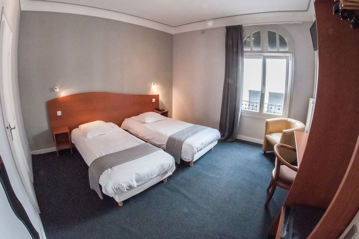 A hotel room with two single beds, a small desk, a chair, and a window with a view.