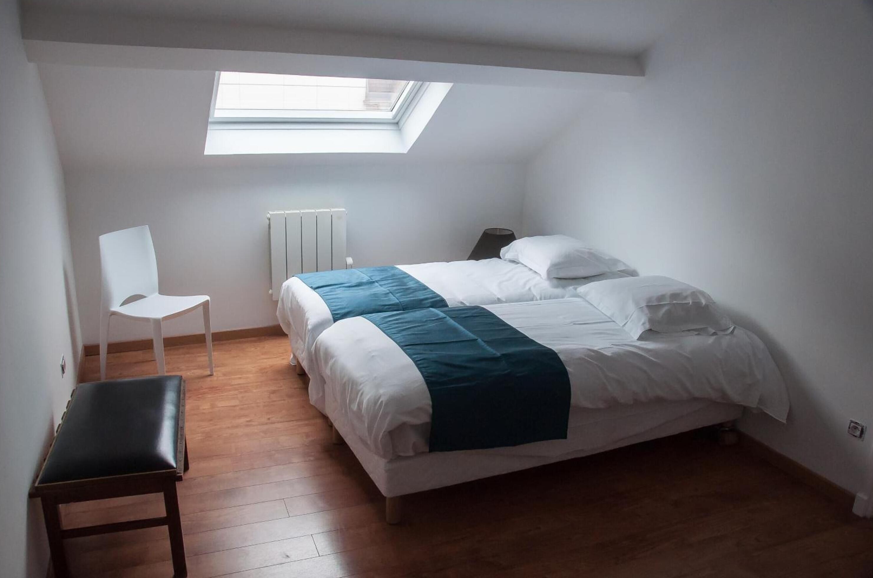 A bedroom with a skylight, a bed with white and teal bedding, a white chair, and a radiator.