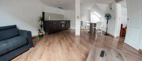 A modern living room with a sofa, a coffee table with a remote, a dining area with chairs, a kitchen with dark cabinets, and a fan.