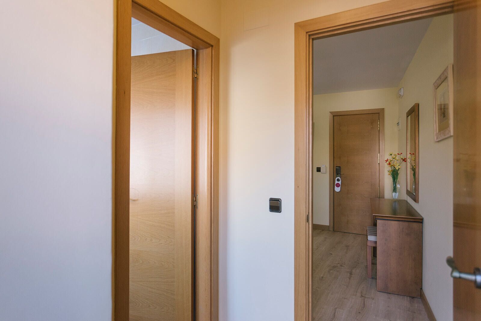 A hallway with wooden doors, a small desk with a vase of flowers, and a mirror on the wall.