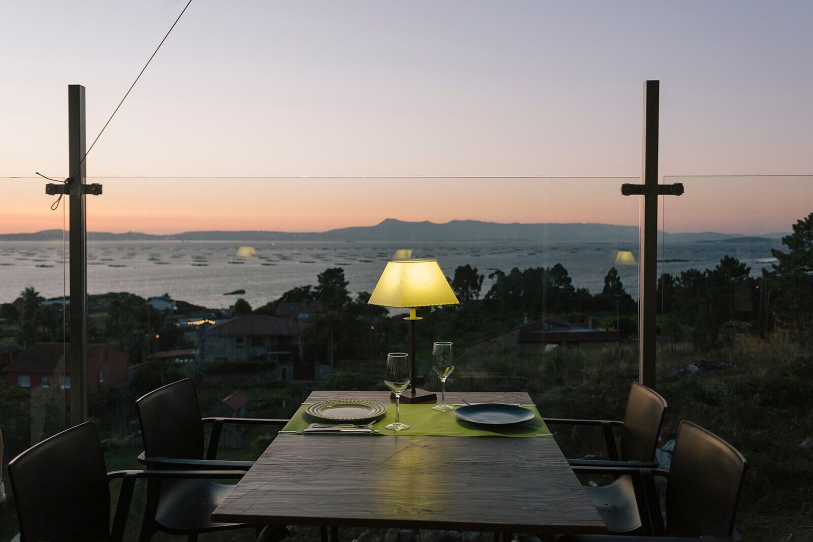 A table set for two with a lamp, glasses, and plates, overlooking a scenic view of the sea and hills.