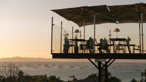 A glass-enclosed outdoor dining area with tables and chairs, overlooking a body of water at sunset. - O Castro (O Grove)