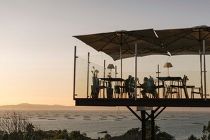 A glass-enclosed outdoor dining area with tables and chairs, overlooking a body of water at sunset. - O Castro (O Grove)