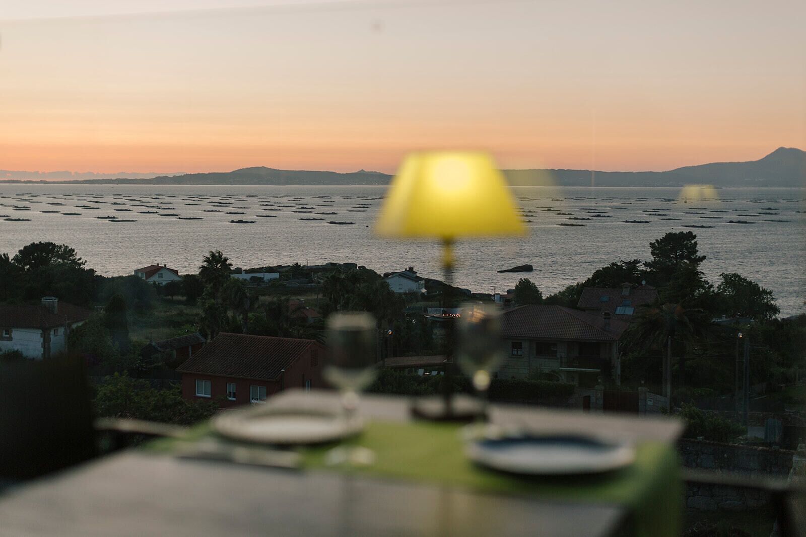 A table with a yellow table lamp, a wine glass, and a plate, overlooking a body of water with boats and a distant mountain range.