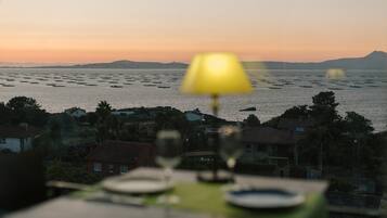 Una mesa con una lámpara de mesa amarilla, una copa de vino y un plato, con vistas a una masa de agua con barcos y una cadena montañosa distante.