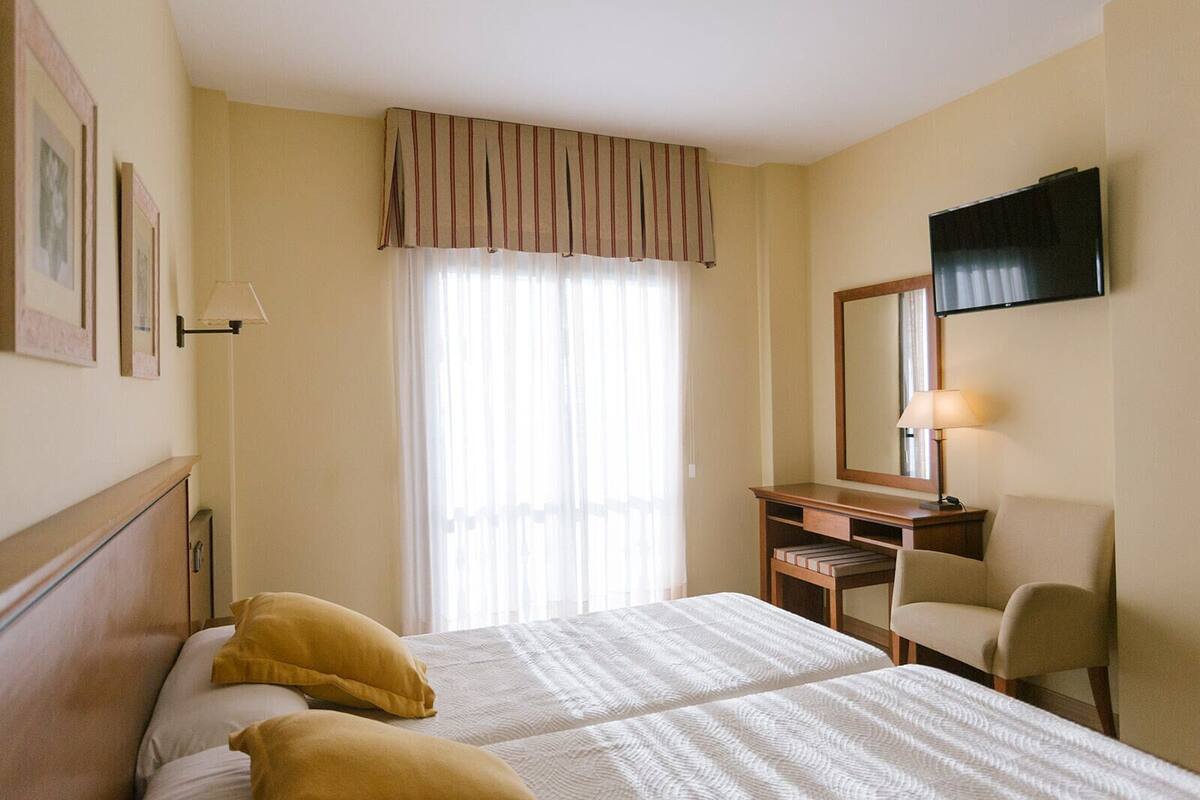 Una habitación de hotel con cama, escritorio, silla, televisor y ventana con cortinas.