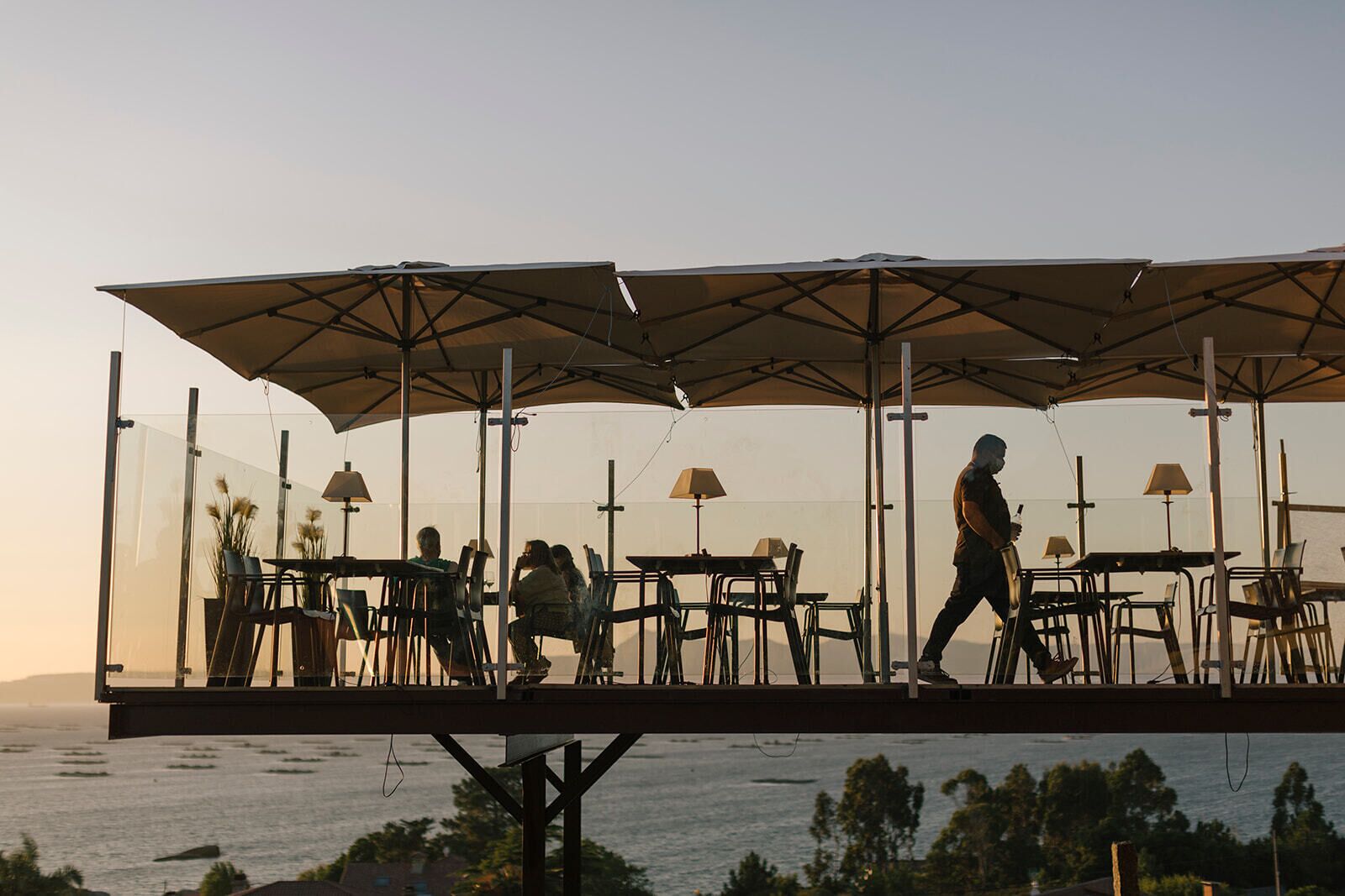 An outdoor dining area with tables and chairs under large umbrellas, overlooking a body of water.