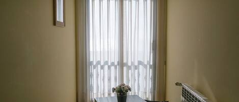 Una habitación estrecha con una mesa, un radiador y una ventana con cortinas traslúcidas.