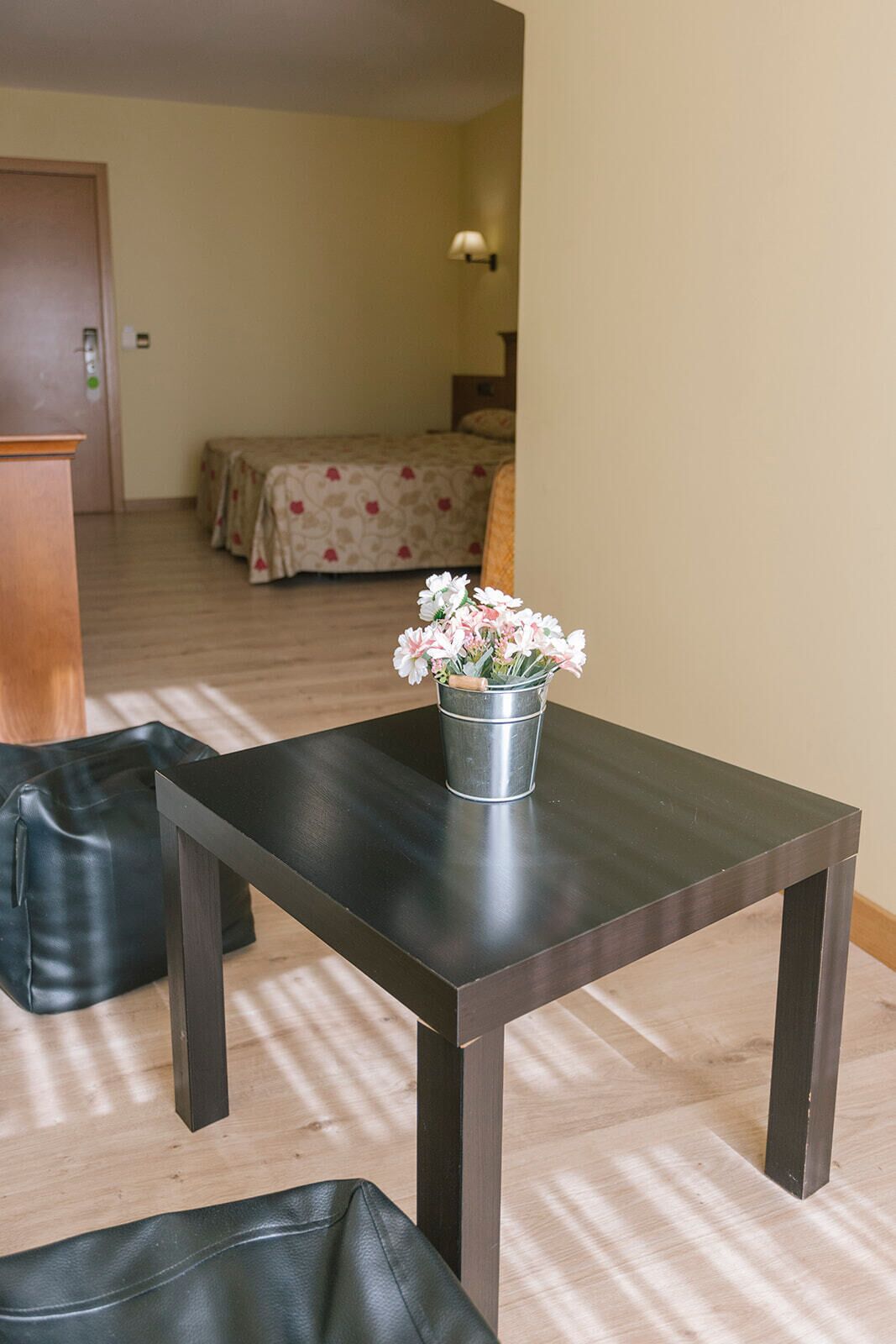 Taula de fusta petita i fosca amb un cubet metàl·lic que conté flors blanques, situada en una habitació amb un llit al fons i un terra de fusta.