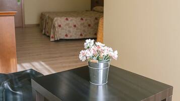 Taula de fusta petita i fosca amb un cubet metàl·lic que conté flors blanques, situada en una habitació amb un llit al fons i un terra de fusta.