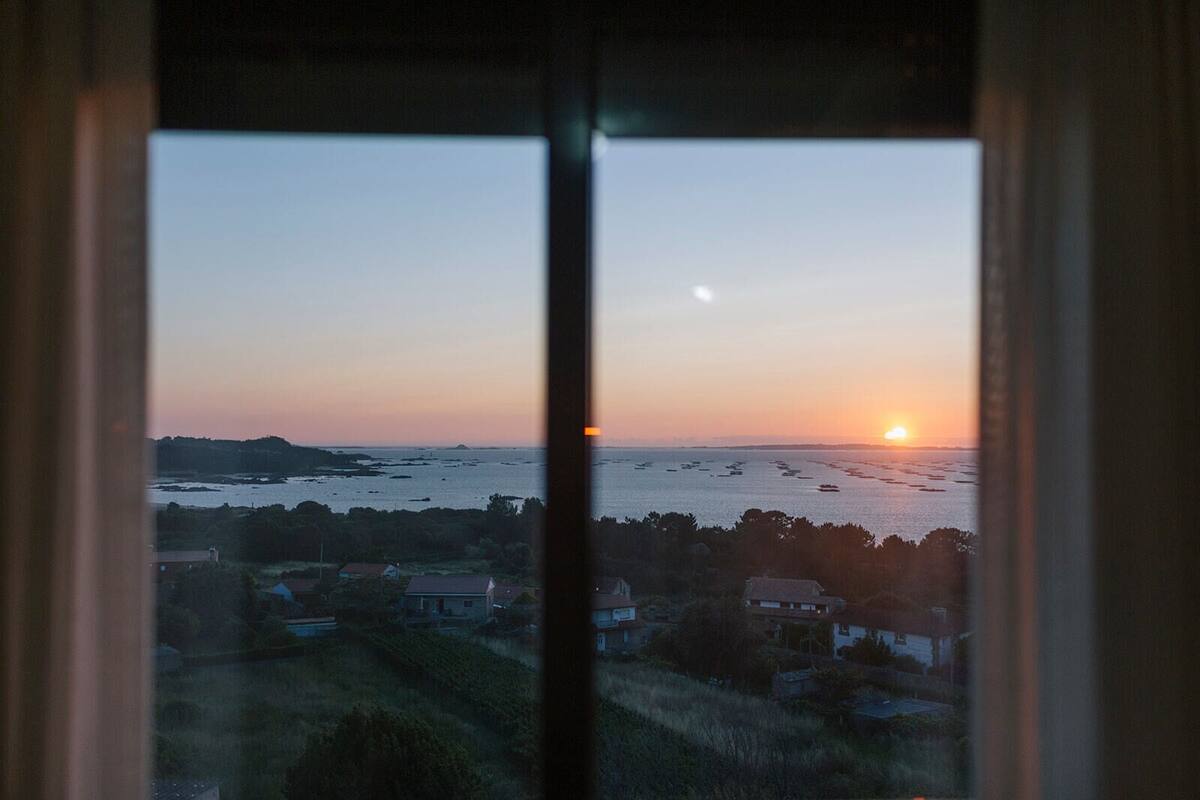Vista a través de una ventana de una zona costera al atardecer, con barcos en el agua y casas en primer plano.