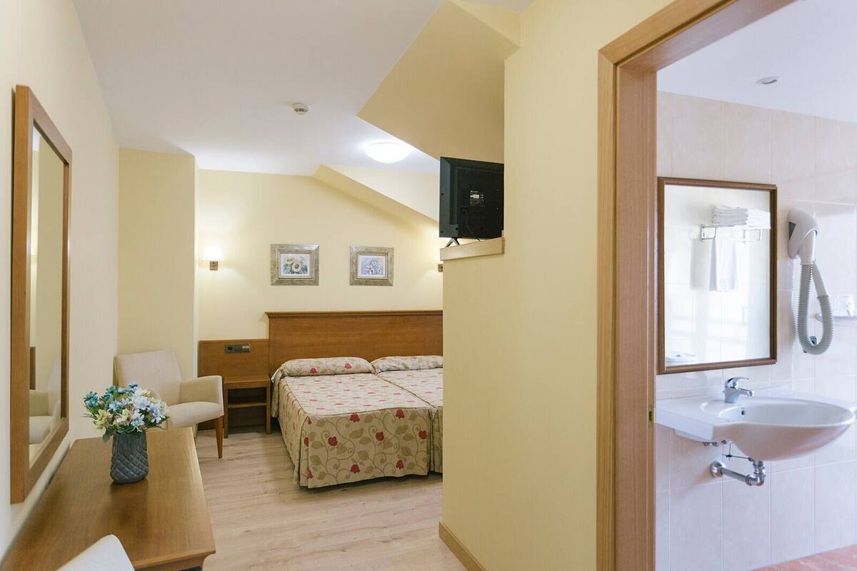 Habitación de hotel con cama, escritorio con un florero, silla, televisor montado en la pared y baño con lavamanos y espejo.