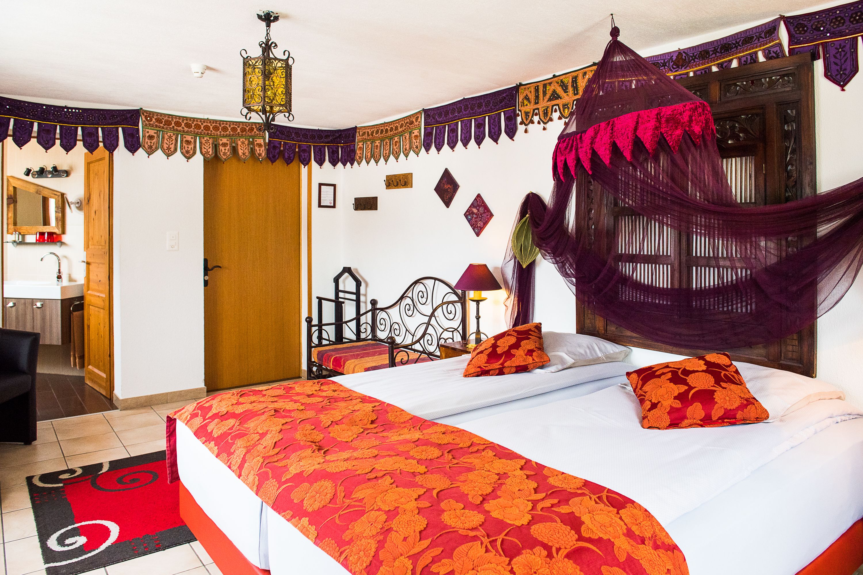 A bedroom with a bed, a nightstand, a chair, a wooden door, a mirror, and a hanging lamp.