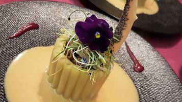 Un postre con una flor morada y brotes sobre un plato texturizado, acompañado de salsa cremosa.