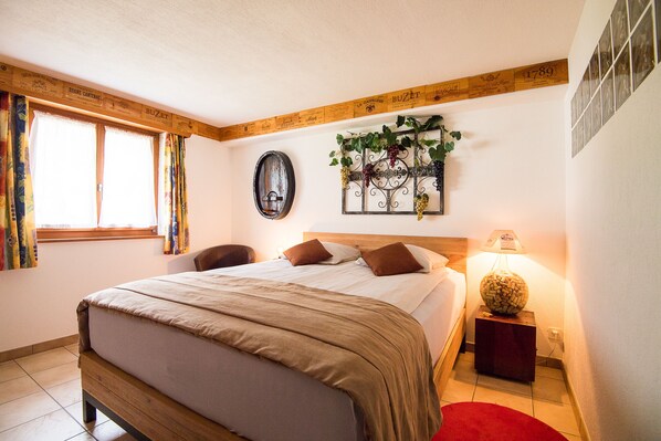 A bedroom with a wooden headboard, a bed with a beige bedspread, two brown pillows, a bedside table with a lamp, and a wall-mounted decoration with grapes. - Rêves Gourmands (Vernayaz)