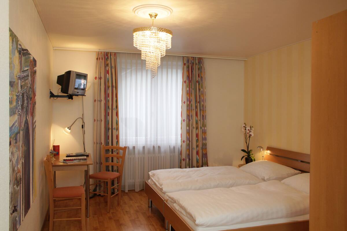 A hotel room with a bed, a desk with a chair, a television, and a chandelier.