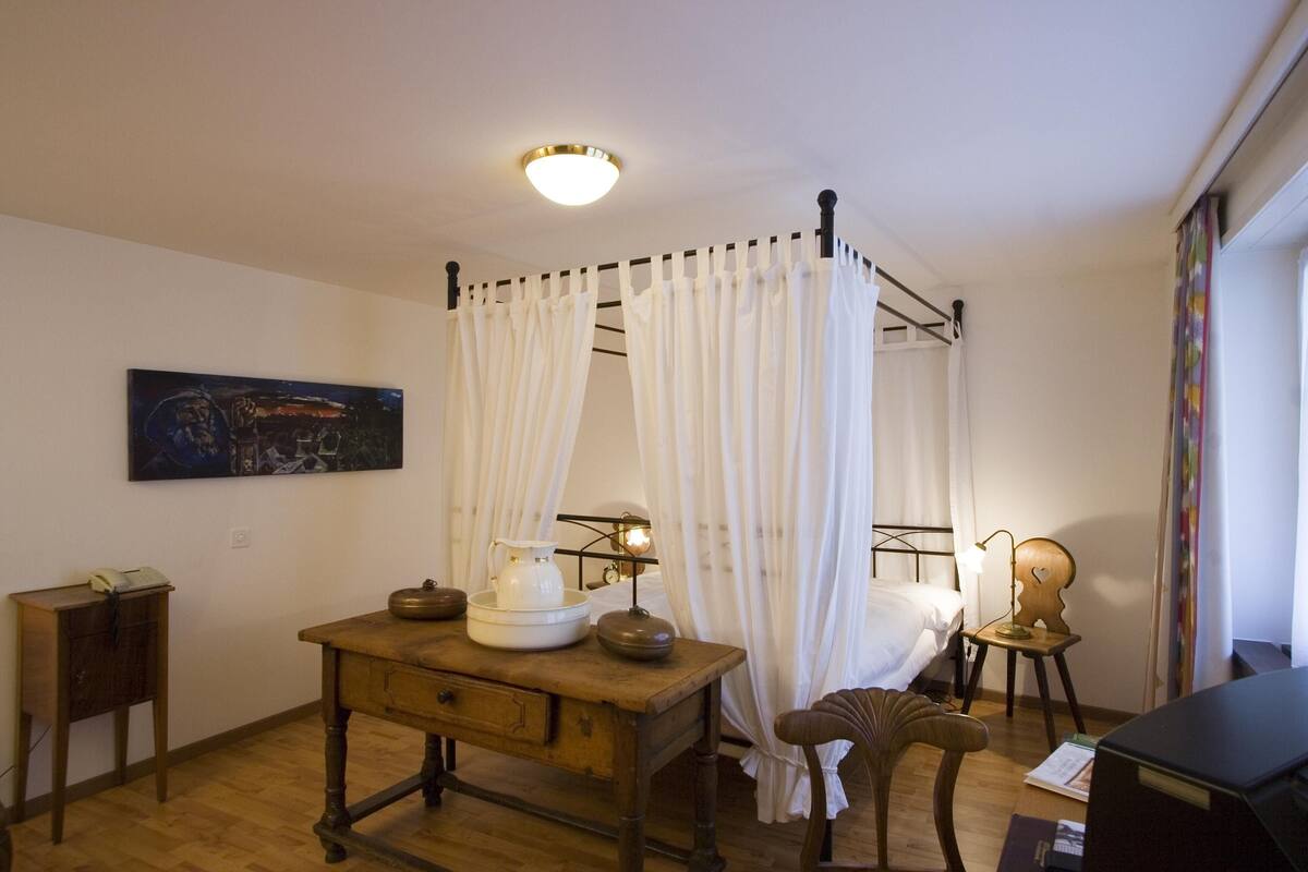 A room with a four-poster bed, a wooden desk with a lamp, a chair, and a painting on the wall.