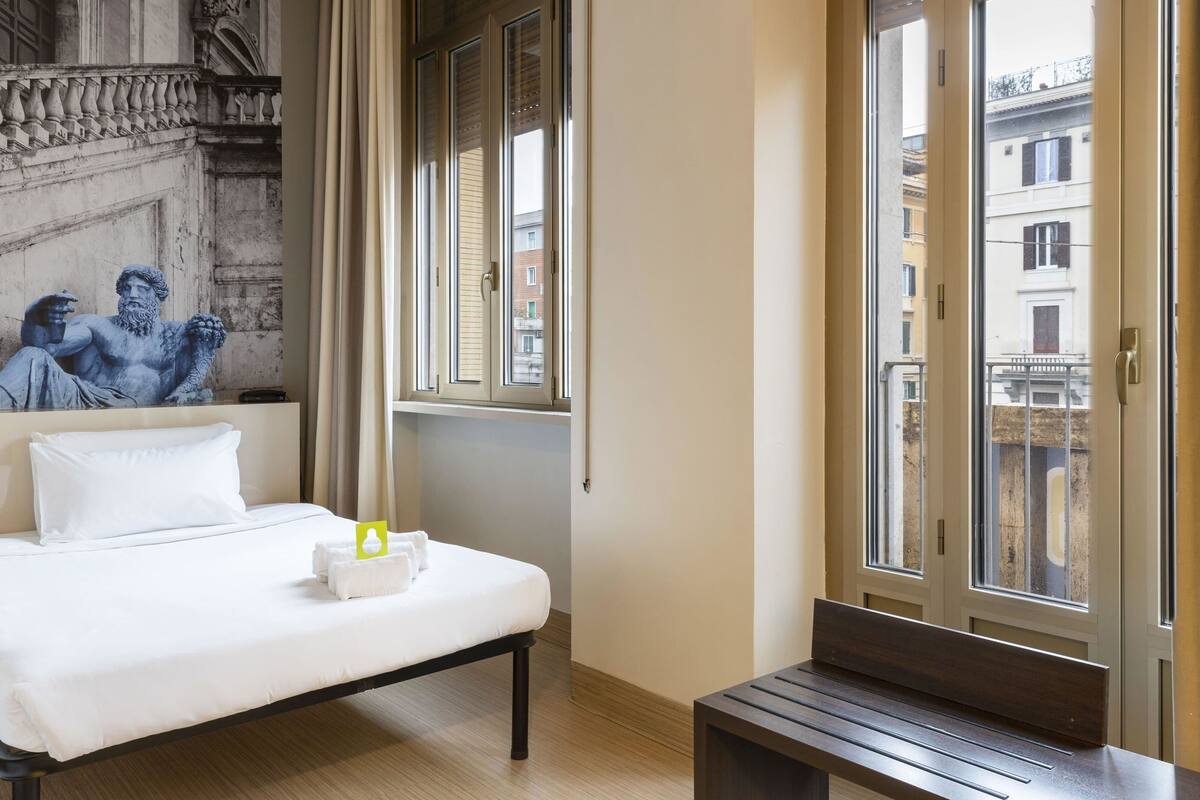 A hotel room with a bed, a bench, and a view of the city through large windows.