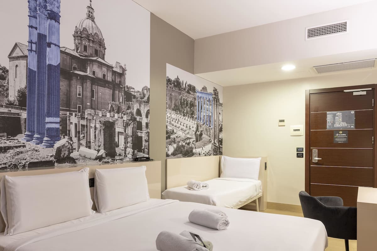 A hotel room with two beds, a desk, and a chair. There are two large wall-mounted artworks depicting historical buildings.