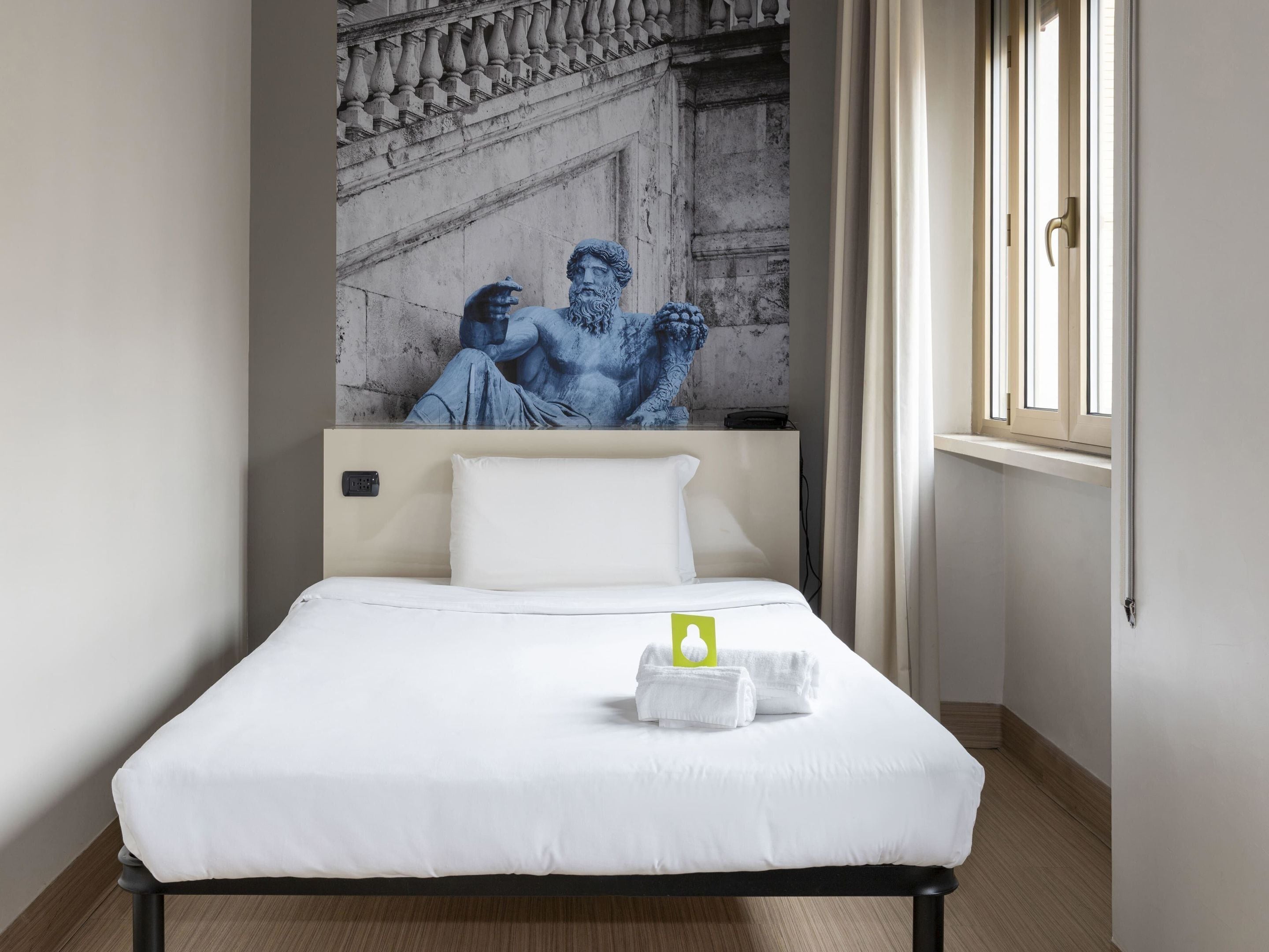 Ein Hotelzimmer mit Bett, weißer Bettwäsche, einem Wandbild einer klassischen Skulptur, einem Fenster mit Vorhängen und einem Nachttisch mit einem grünen Gegenstand.
