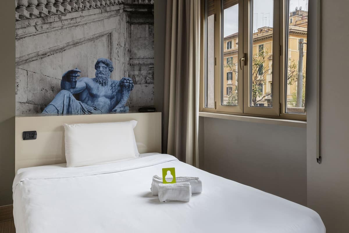 A hotel room with a bed, a painting of a classical sculpture on the wall, and a window with a view of buildings.