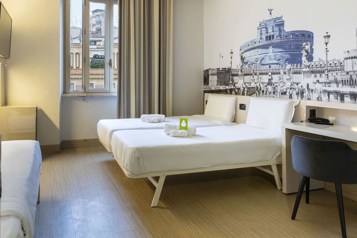 A hotel room with two beds, a desk, a chair, a window with curtains, and a wall-mounted TV.