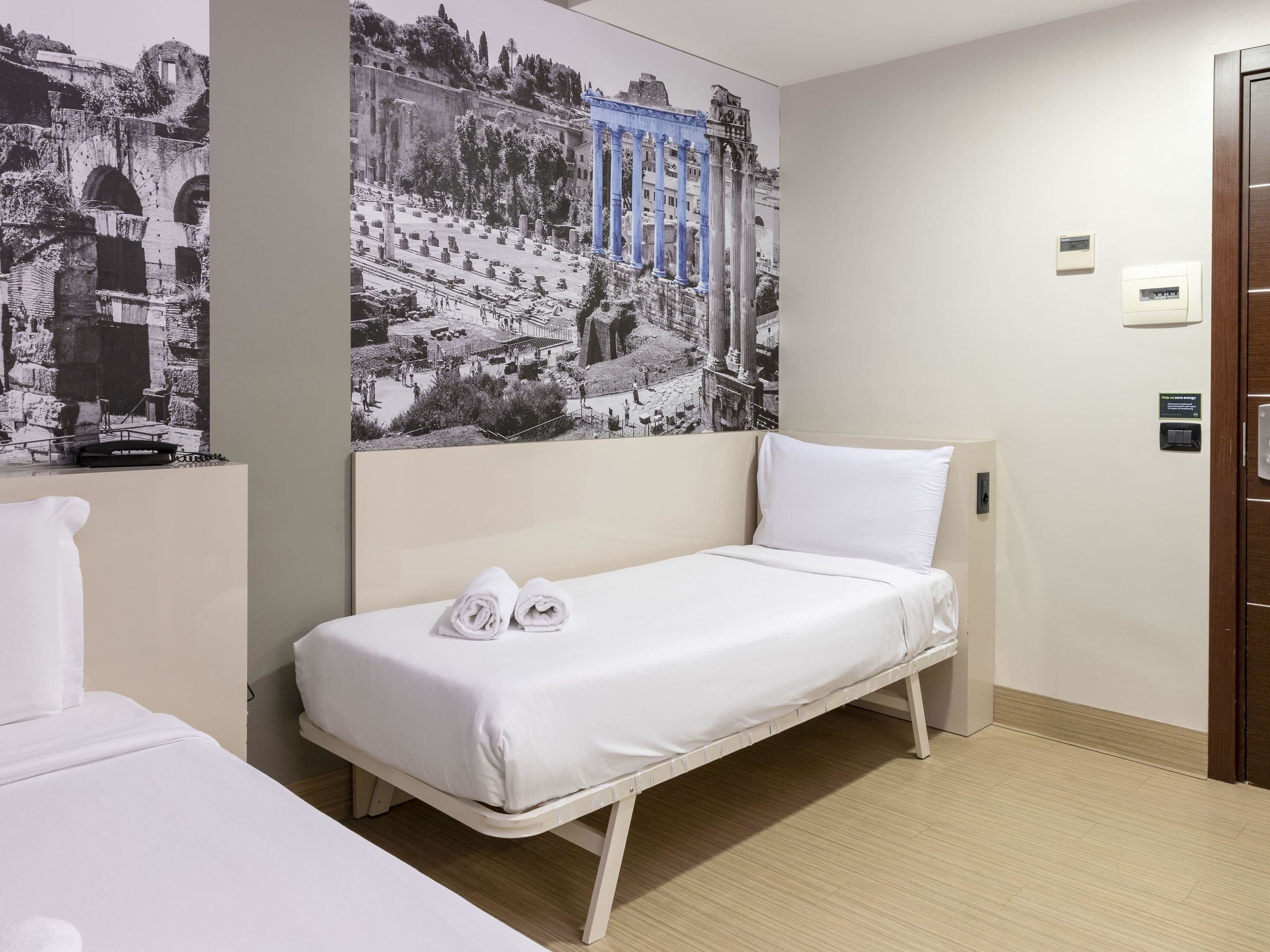 Een eenpersoonskamer met een wit bed, hoofdeinde en een kunstwerk aan de muur dat een oud stadsgezicht toont.