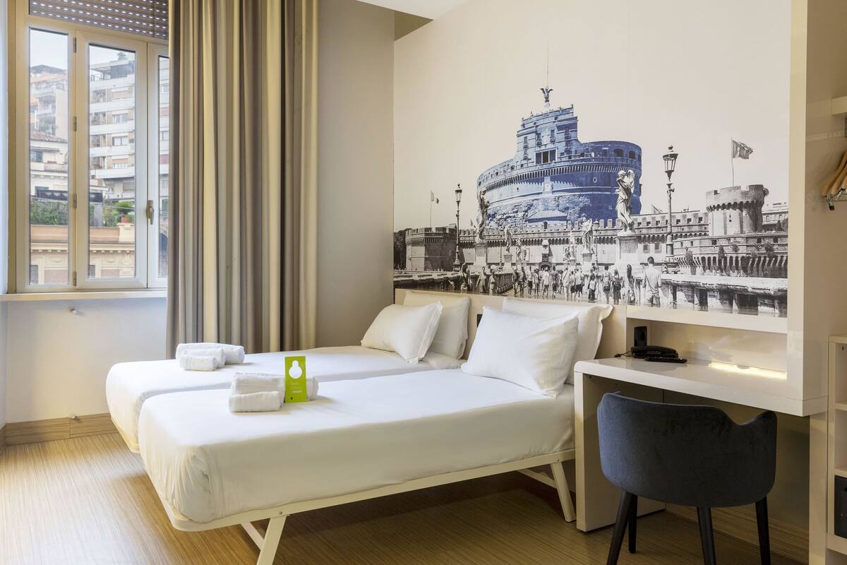 A hotel room with two beds, a desk, and a chair. There is a large window with curtains, and a wall mural depicting a historical building.