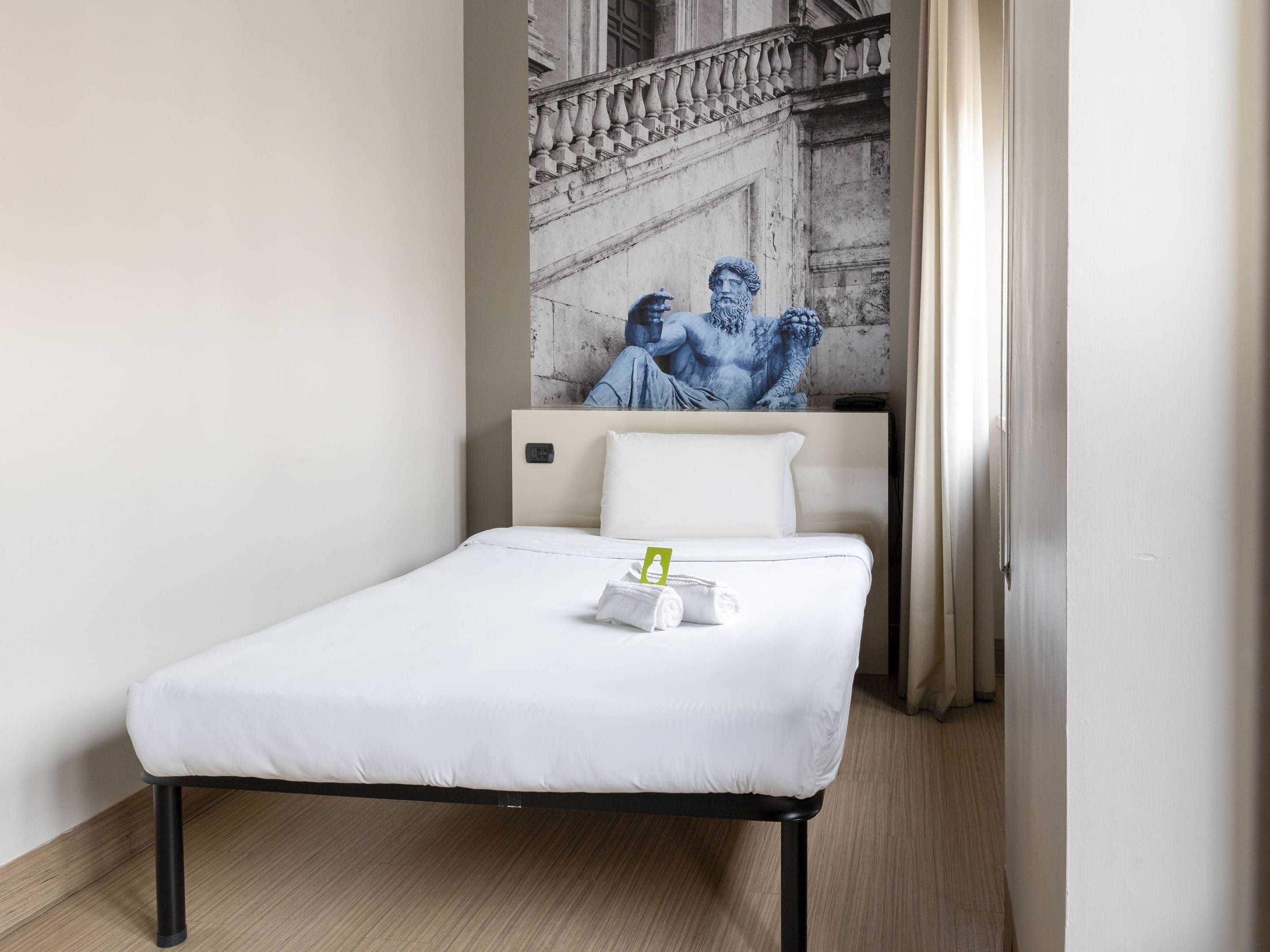 A single bed with a white mattress and black frame, a small bedside table with a green object, and a wall-mounted artwork of a classical sculpture.