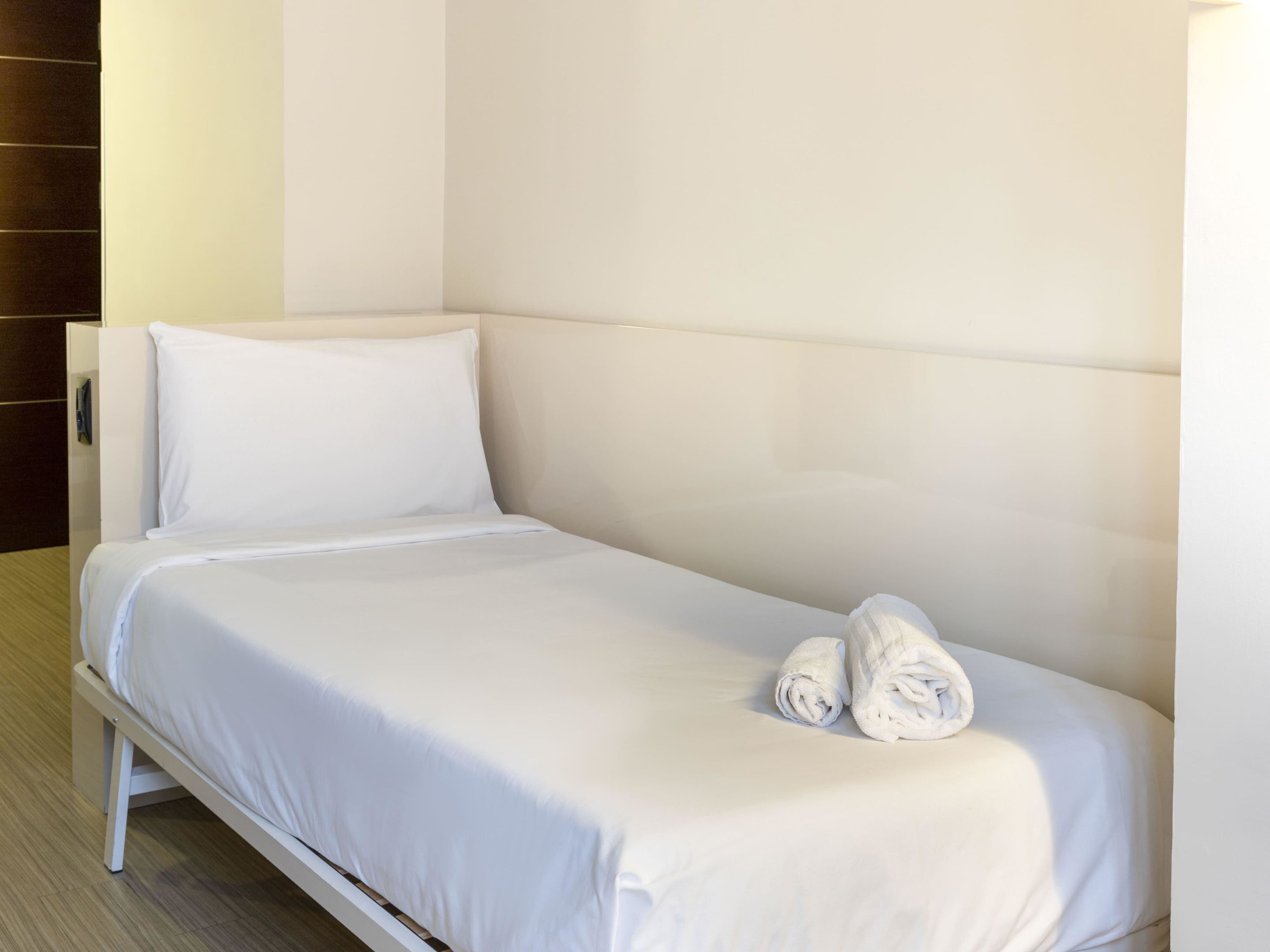 A small, neatly made bed with white linens and two rolled towels on the side.