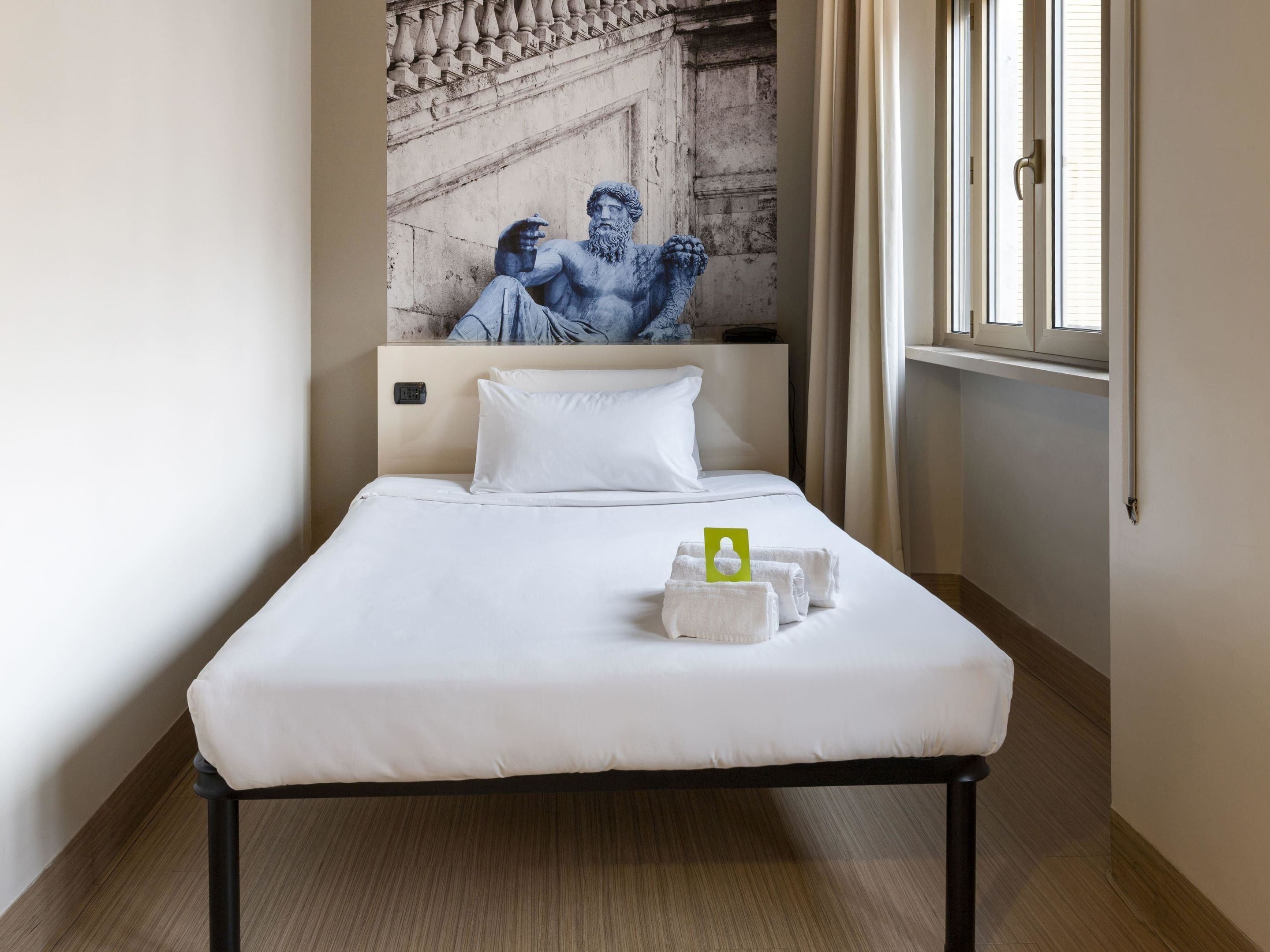 Ein Hotelzimmer mit einem Bett, einem Bild einer klassischen Skulptur, einem Fenster und einem Handtuchhalter mit Handtüchern.