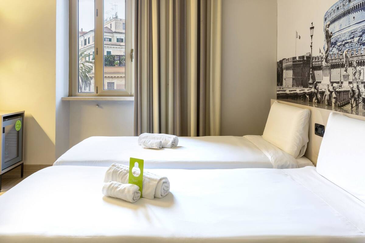 A hotel room with two beds, a window with curtains, a wall with a historical mural, and a small table with a lamp.